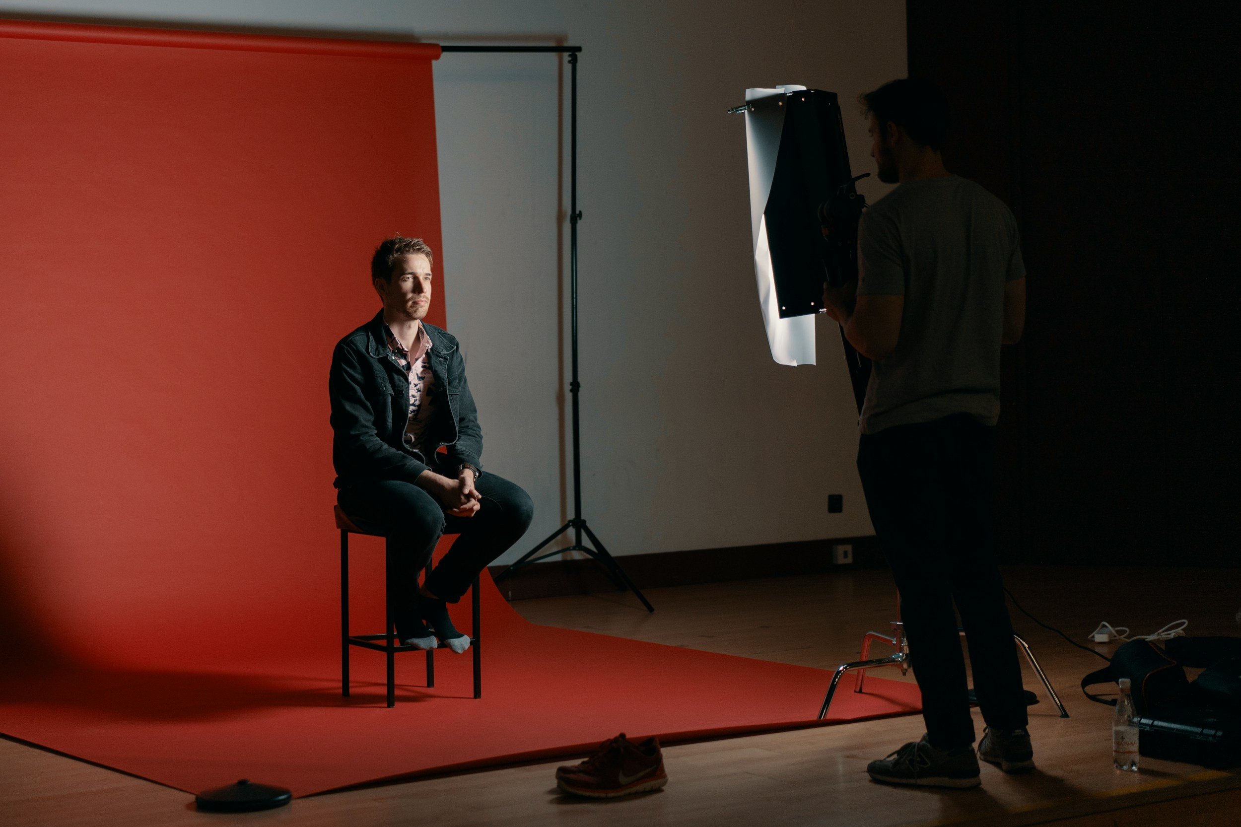 An image of a person sitting on a chair with a red backdrop behind them. A Lighting Camera Operator is lighting the scene.