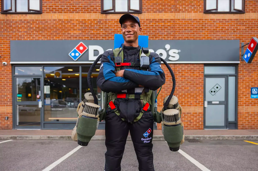 A pilot from gravity industries, wearing a jetpack, in front of a dominos store.