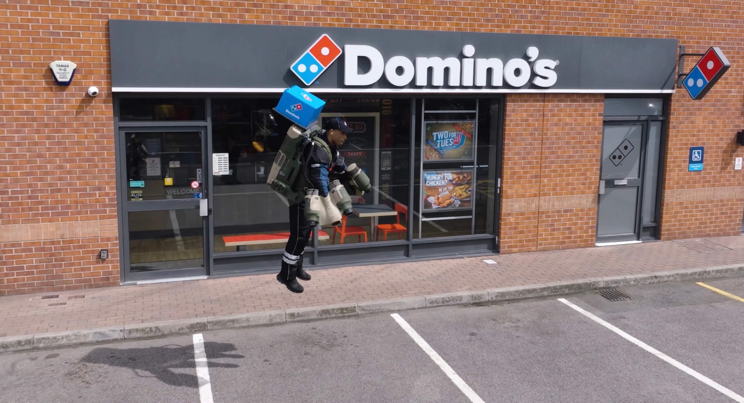 A drone shot of the outside of a Dominos shot as a jetpack pilot takes off with a Pizza.