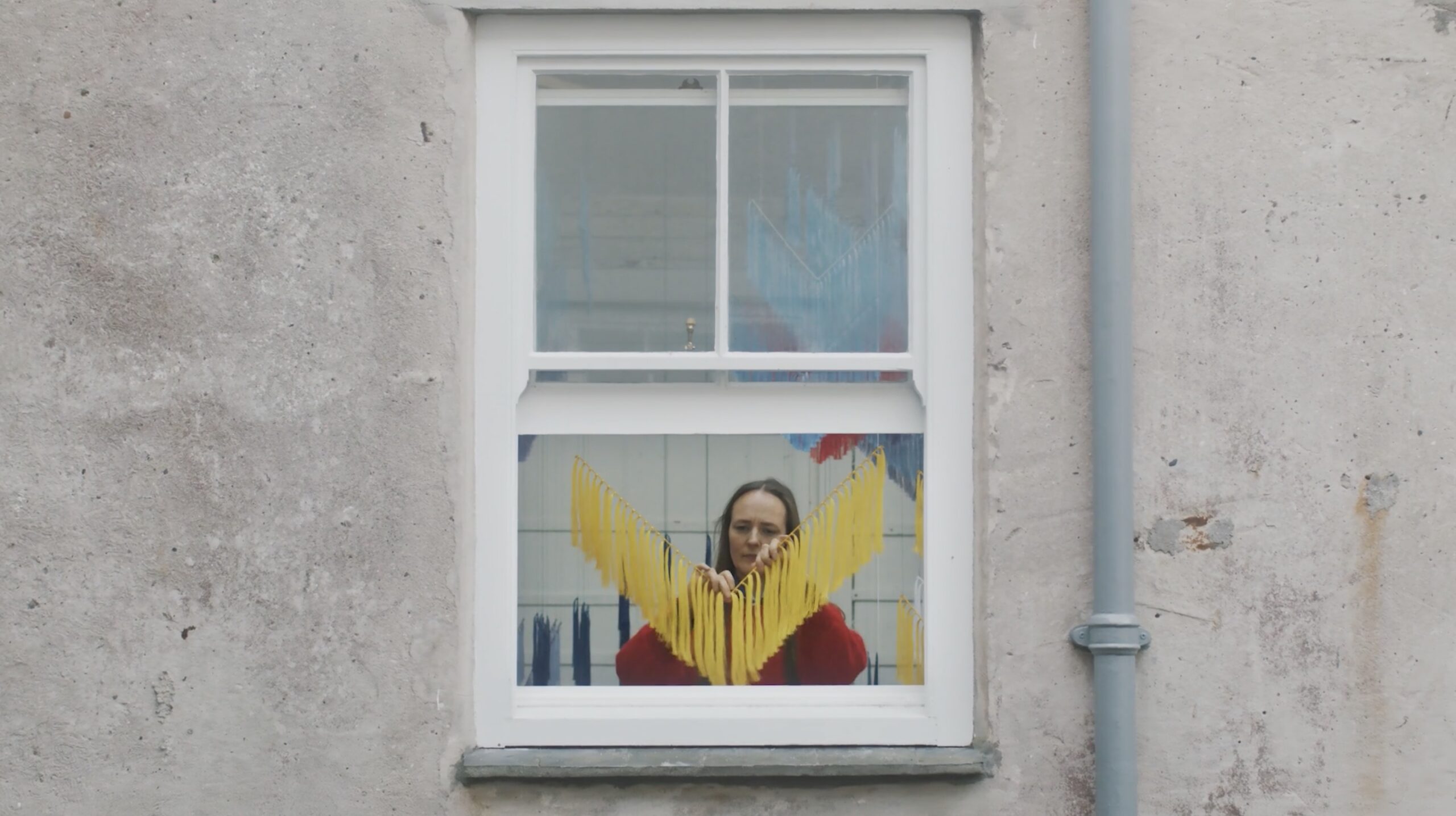 An image of Outi Pieski making her most recent piece of art for The tate. The image is taken through an open window showing the outside of the studio.