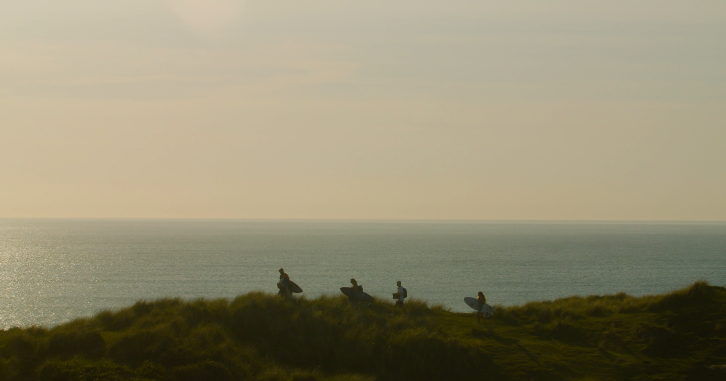 An image of 5 people walking through sand dunes, silhouetted with the sea behind them.