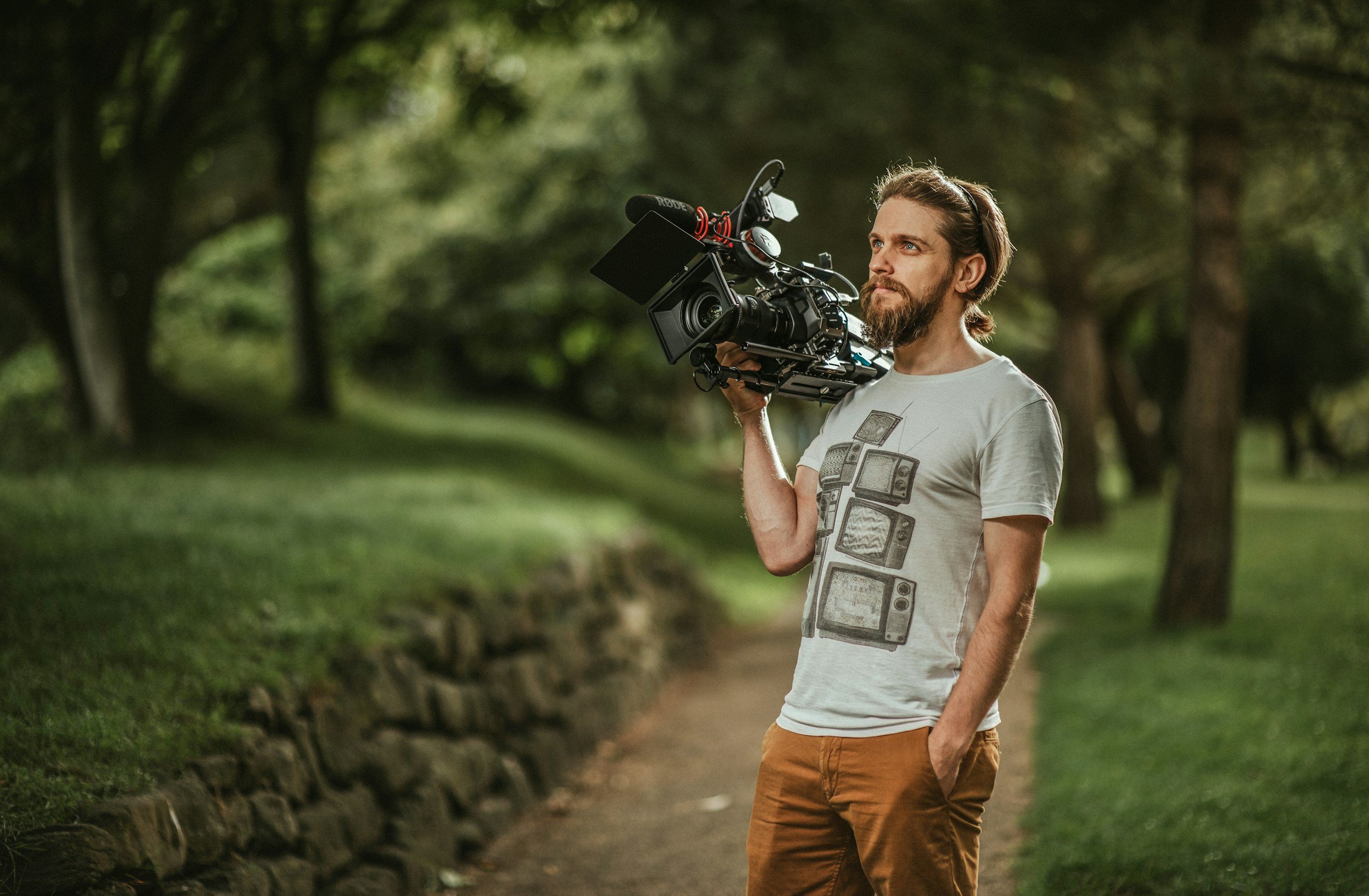 Male camera operator with brown hair with a camera on his shoulder. Trees and garden are in the background.