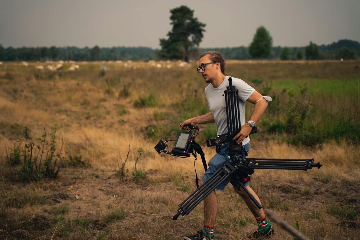 A camera assistant carrying a tripod across a field.