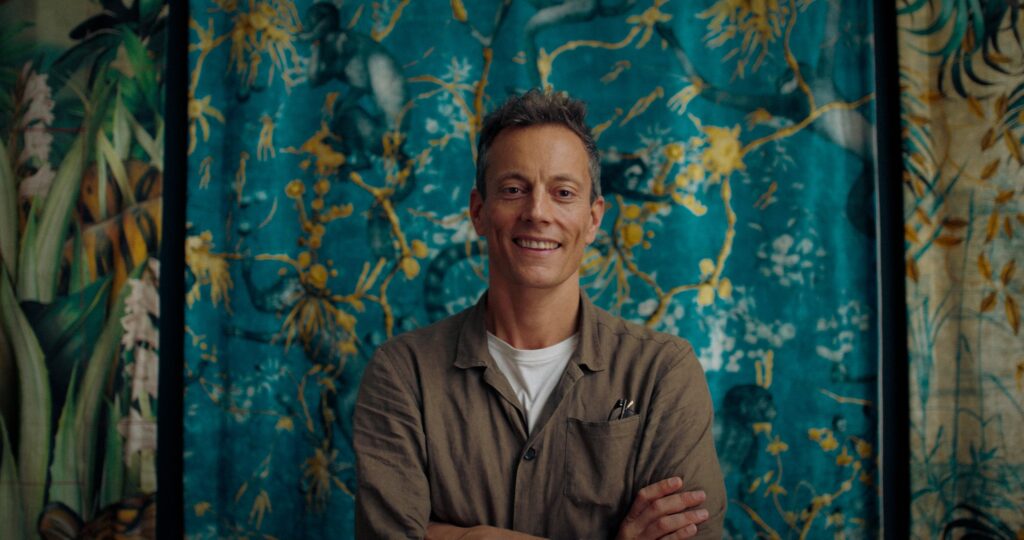 An image of Adam Elis, designer based in London, smiling into the barrel of the lens with a green tapestry behind him.