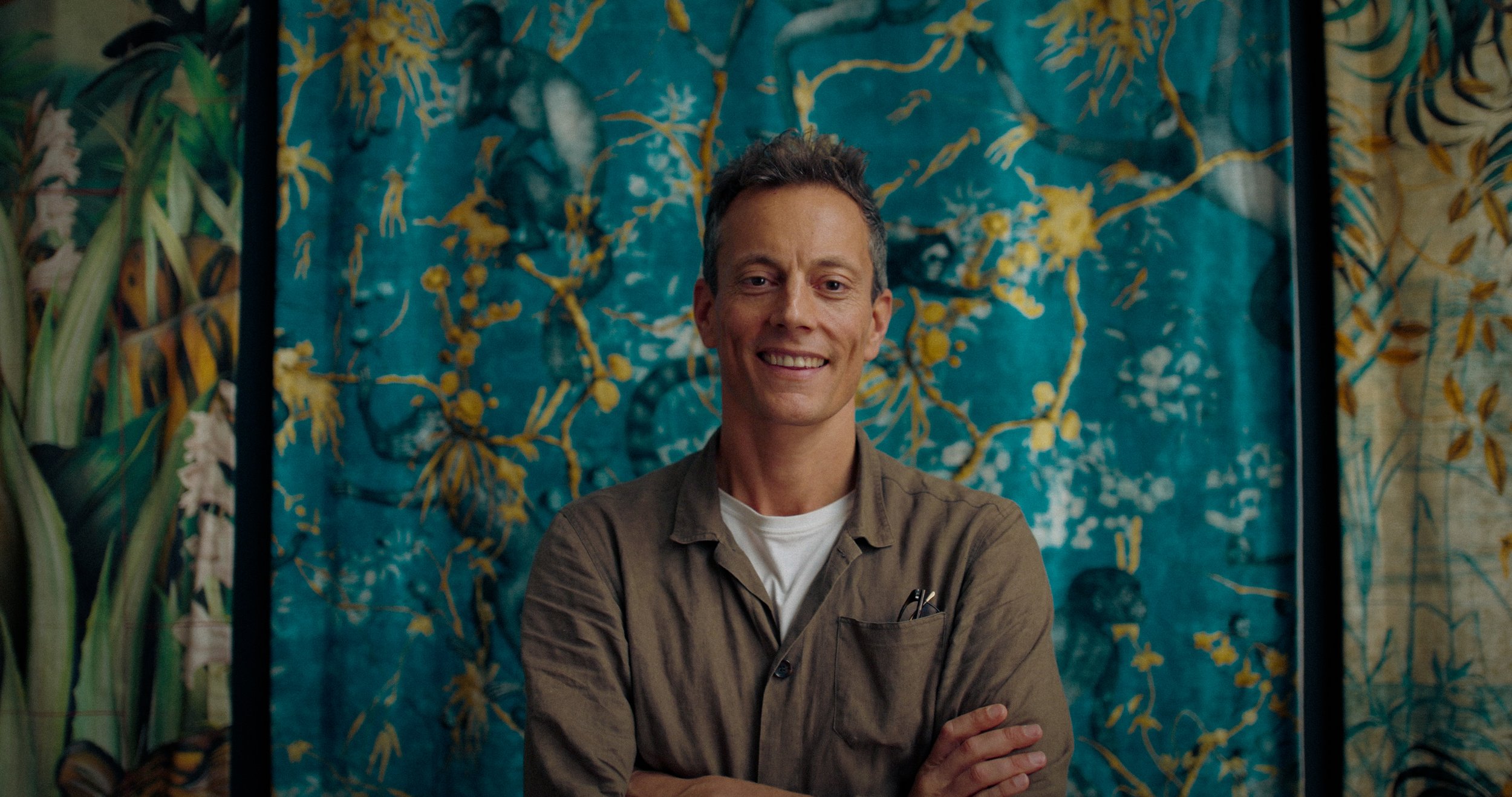 An image of Adam Elis, designer based in London, smiling into the barrel of the lens with a green tapestry behind him.