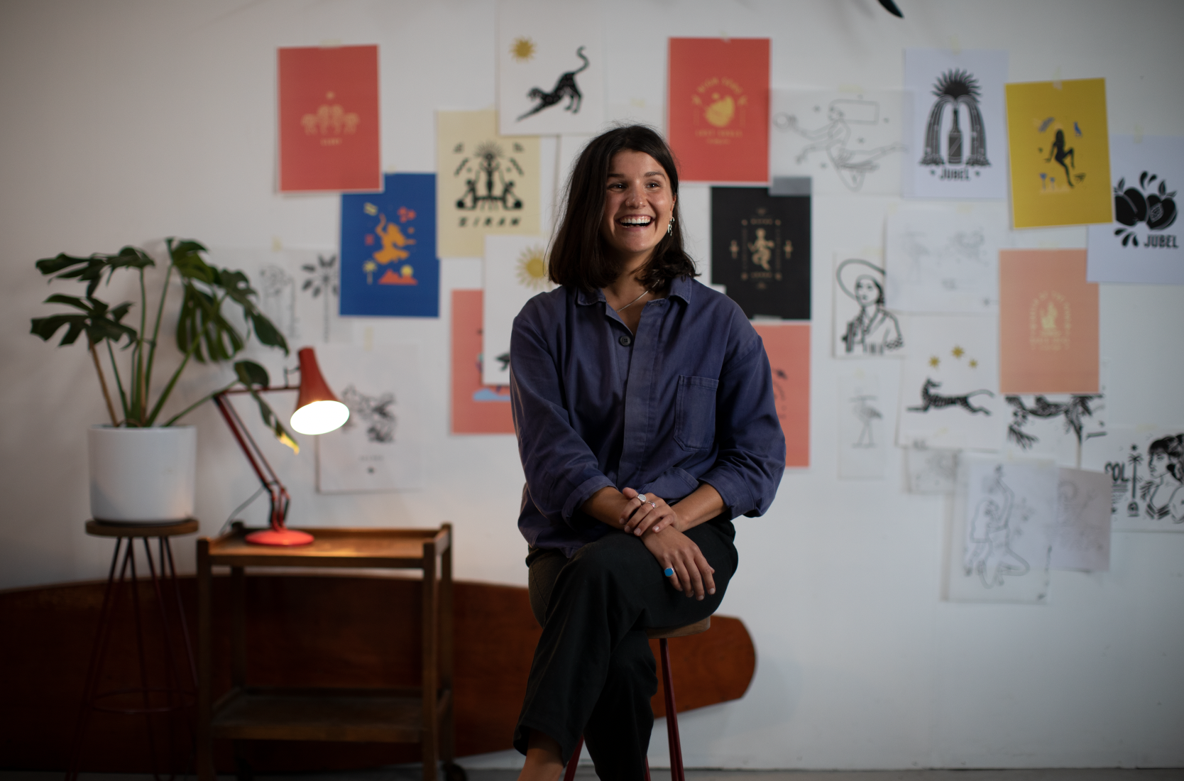 Clara Jonas sitting on an interview chair in front of a wall of her work.