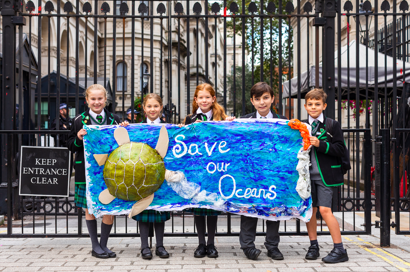 A photo of school children holding up a banner by the gates of downing street. the sign says "Save the ocean".