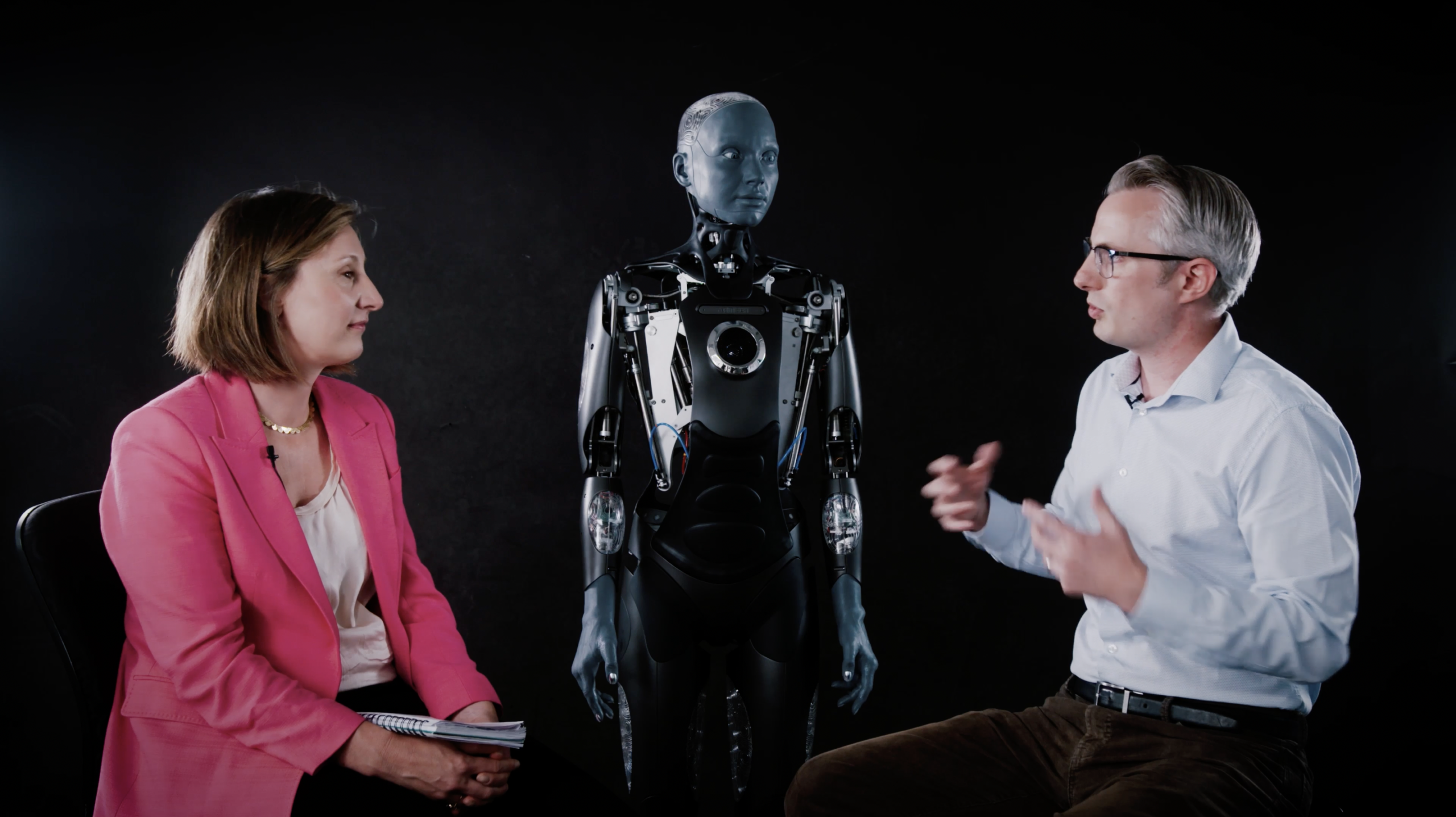 An image of two perople speaking in front of AMECA the humanoid robot.